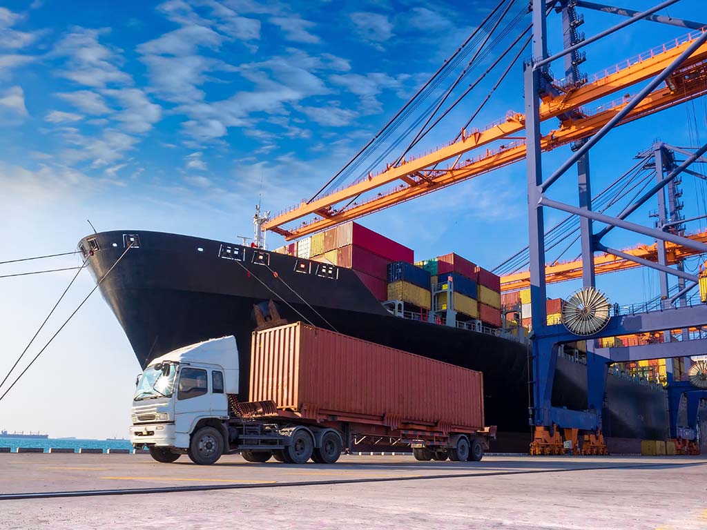 A shipping container on a truck in front of a cargo ship